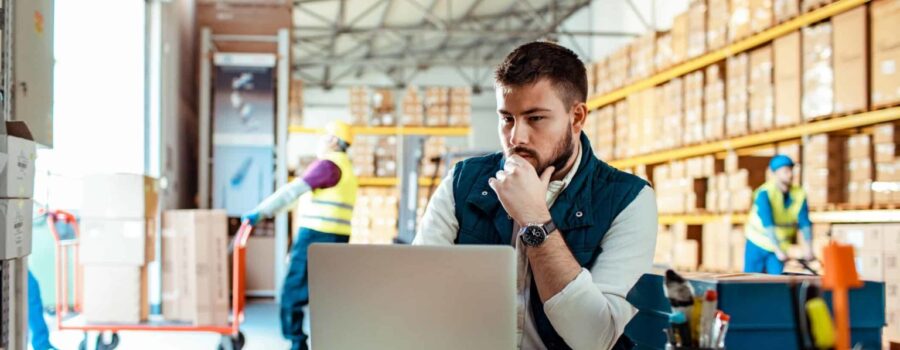 A man focuses on a laptop in a warehouse, with shelves of boxes and two workers organizing stock in the background.