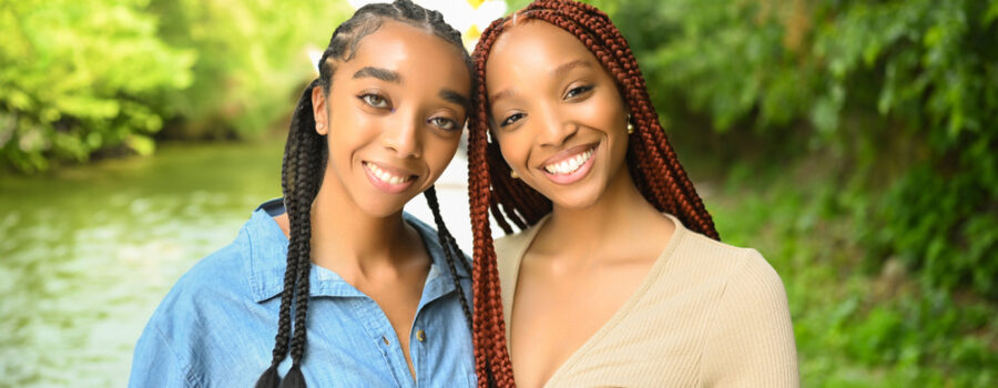 Two women standing outdoors near a river, smiling at the camera. One has long braids and wears a blue shirt, the other has red braids and wears a beige top. Greenery surrounds the background, capturing a serene moment as they embark on their exciting journey to study in the USA.