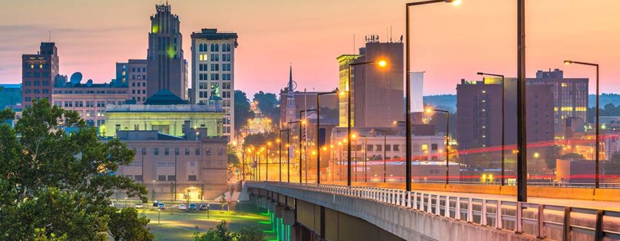 A cityscape at dusk featuring a bridge leading into the downtown area with illuminated buildings and streetlights glowing against a colorful sky.