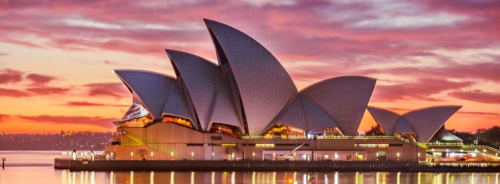Sydney Opera House with its distinct sail-like design, illuminated during sunset, reflected in the water.