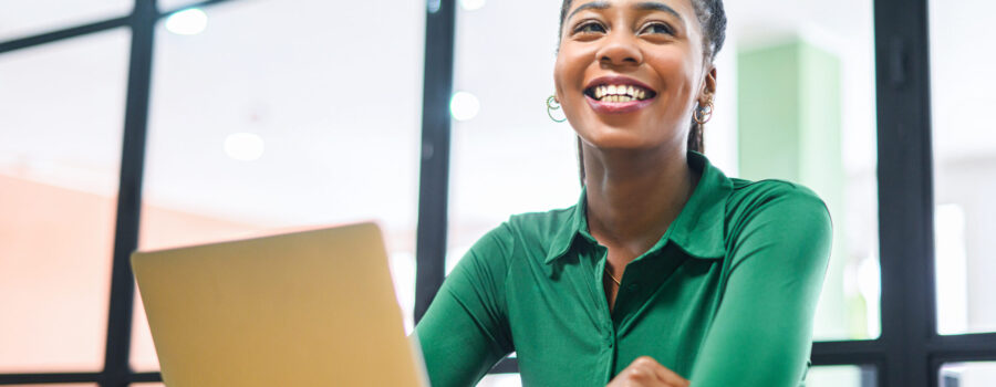 A person in a green shirt sits at a desk with an open laptop, smiling and looking slightly upward. A glass window in the background reveals the blurred office setting where work permits are being processed.