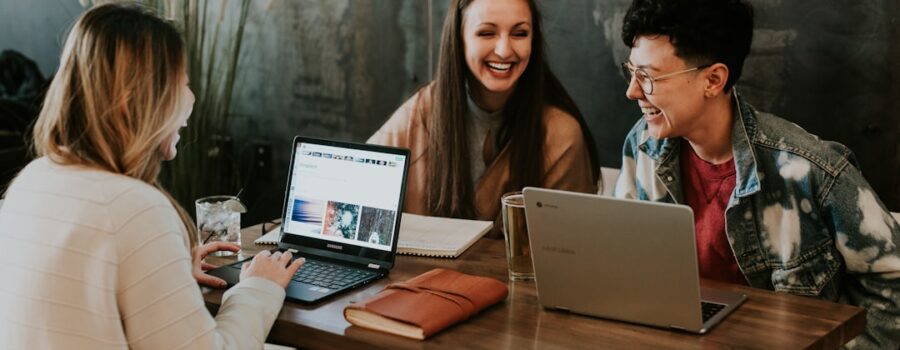 Three people sitting at a table with laptops, smiling and talking. A book and a glass are on the table. They are discussing their plans to study in the United States in a casual environment with a concrete wall and a plant in the background.