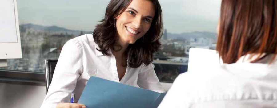 A woman in a white shirt holding a blue folder smiles while conversing with another person in an office setting, possibly preparing for her U.S. Visa interview.