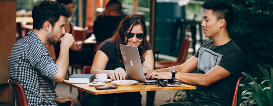 Three international students sit around a table at an outdoor café, engaged with a laptop. Coffee cups and notebooks are scattered across the table as they exchange ideas and embrace their diverse perspectives.