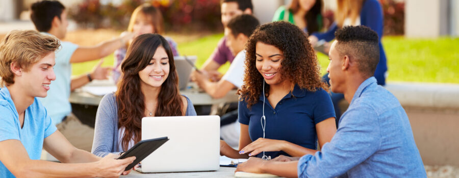 A group of students living in the UK sits at an outdoor table, using a laptop and tablet while conversing. Other groups of students are visible in the background.