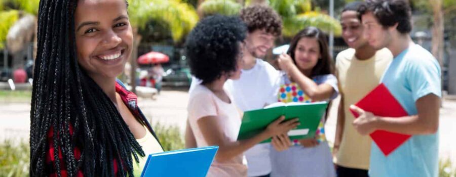 A smiling person holding a blue folder stands in the foreground, while a group of five people with books and folders engage in conversation about work in the UK in the background outdoors.