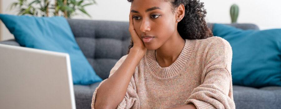 A person with curly hair sits at a desk, looking at a laptop screen with a thoughtful expression. The background features a grey sofa and blue cushions.