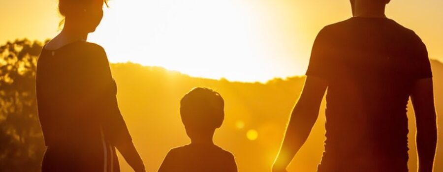 Silhouetted view of two adults and a child holding hands, facing a bright sunset.