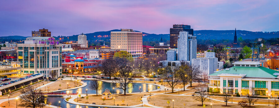 Aerial view of a cityscape at dusk, featuring water bodies, city buildings, and a backdrop of hills under a colorful sky.