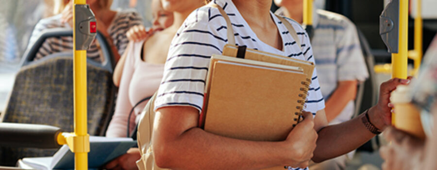 A woman standing on a bus, holding notebooks, and smiling. Other passengers are seated in the background.