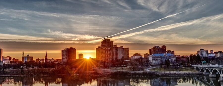 City skyline at sunrise, with buildings reflecting in a calm river and scattered clouds enhancing the warm, colorful sky.