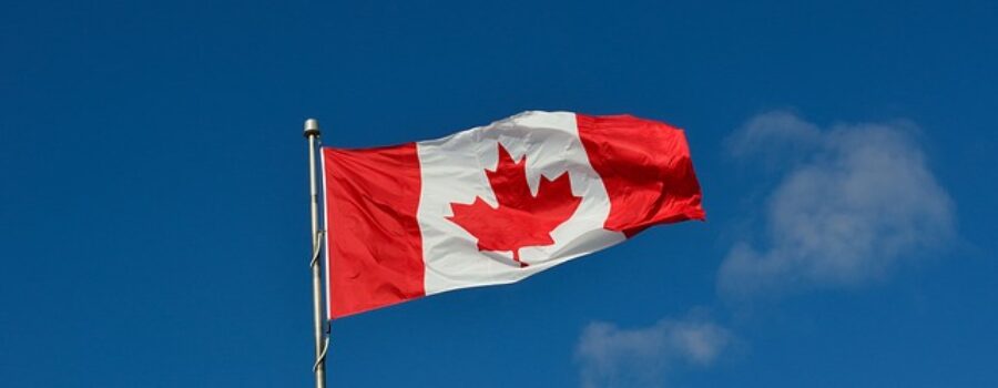 Canadian flag with a red maple leaf centered on white, flanked by red vertical stripes, waving against a clear blue sky.