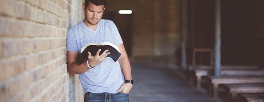 Man in casual clothing leans against a brick wall, reading a book in a dimly lit corridor.