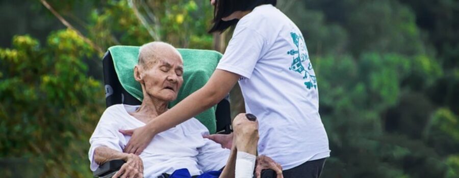 A person assists an elderly man seated in a wheelchair outdoors, with trees in the background.