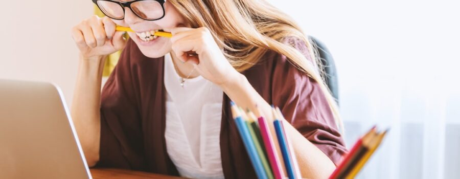 A woman with glasses bites a pencil while looking at a laptop screen. A cup of colorful pencils is on the desk.