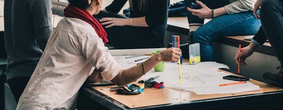 Four people sit and lean around a table with papers, pens, and a calculator, engaged in discussion or group work in a casual indoor setting.