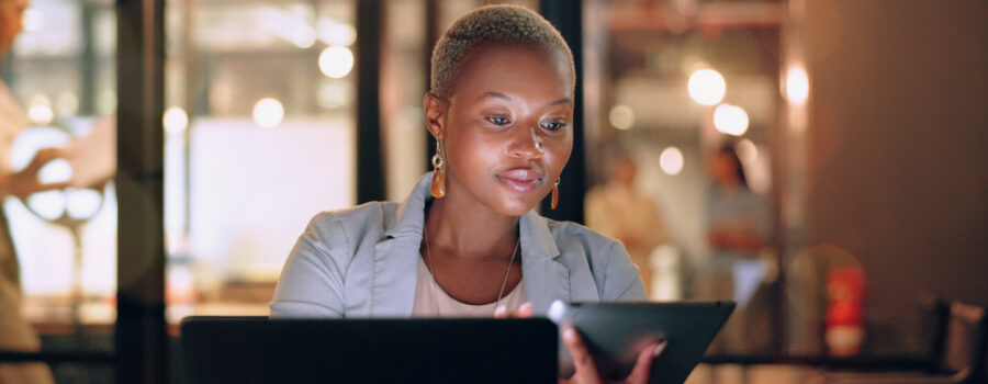 A person sits at a desk in an office, working on a tablet and a laptop, with blurred lights and people in the background.